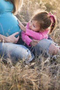 Защита от домашнего насилия (беременная женщина и ее маленькая дочка сидят на корточках на траве и девочка гладит мамин живот)