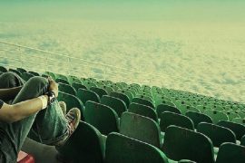 Одинокий человек на трибуне стадиона, поле которого символически изображено, как бесконечное пустое пространство. Что делать одиноким людям?