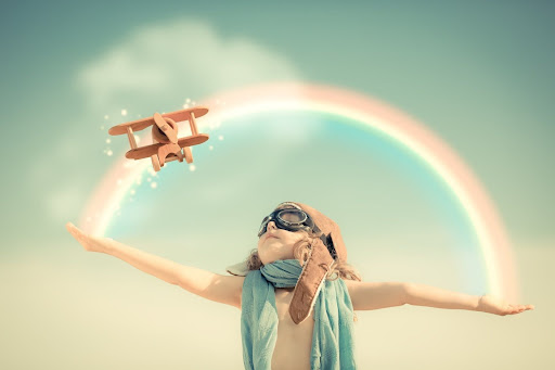 Мальчик в шлеме и очках летчика запускает игрушечный самолетик на фоне чистого неба с радугой. Ребенок, играя, чувствует мир совершенны