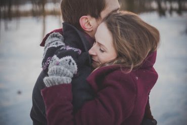 В зимнем парке женщина нежно обнимает мужчину. Объятия повышают уровень гормона любви.