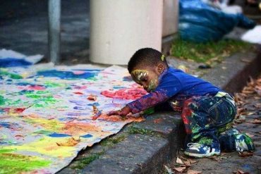 Маленький чернокожий мальчик расписывает лежащую на асфальте бумагу, создает свой художественный образ.