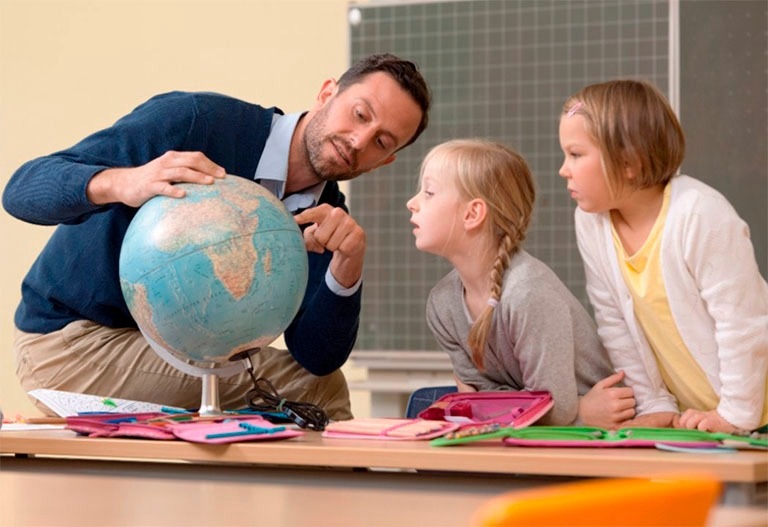 Учитель что-то показывает двум школьницам на учебном глобусе. Учитель должен быть таким, чтобы с ним всегда было интересно.